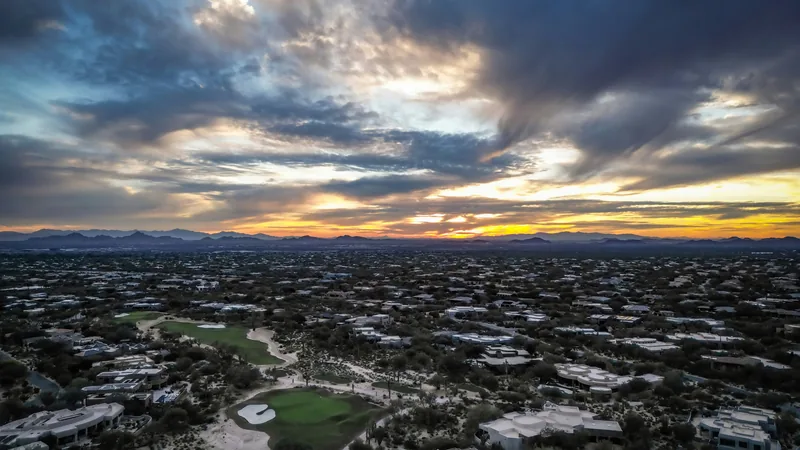 Aerial view of homes and a green fairway among desert trees under a dramatic sunset sky